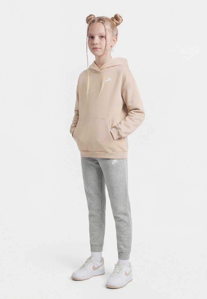 Młoda dziewczyna stoi z rękami w kieszeniach, ubrana w beżową bluzę Nike z kapturem, szare spodnie dresowe Nike i białe sneakersy Nike, na białym tle.