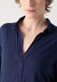 Mornarsko plava košulja s kragnom, V-izrezom i mekanim materijalom. Kragna ima blagu teksturu i leži ravno uz izrez.