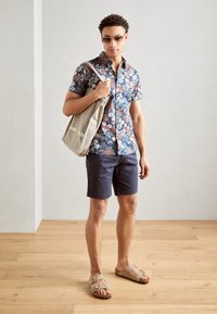 Blomstret kortærmet skjorte i marineblå med pink og blå mønstre, mørke shorts, sandaler med to stropper og en lys farvet totebag.