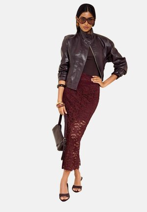 Frau trägt dunkelbraune Lederjacke, burgunderfarbenen Spitzenrock, schwarzes Oberteil, schwarze Absätze, Sonnenbrille und hält eine braune Handtasche.