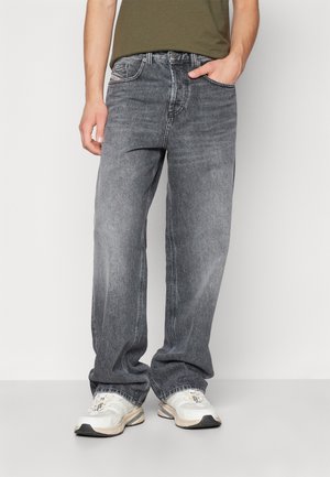Person trägt ausgewaschene schwarze Jeans mit lockerem Schnitt und weiße Turnschuhe und steht vor einem einfarbigen hellen Hintergrund.