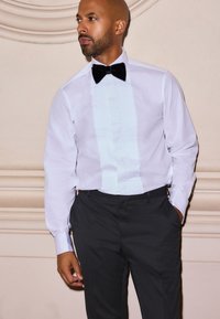 Camicia da smoking bianca con pettorina plissettata, abbinata a pantaloni neri. Presenta un papillon in velluto. Tessuto liscio e design classico formale.