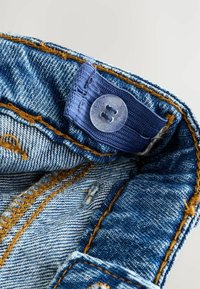 Gros plan sur un tissu en denim montrant une couleur bleu délavé, des coutures oranges et un bouton en plastique clair avec deux trous sur une ceinture.