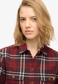 Rood en marineblauw geruite flanellen shirt met korte mouwen, knoopsluiting aan de voorkant, twee borstzakken en goudkleurig logo-detail. Zachte textuur.