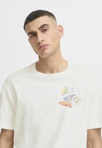 Bomuld T-shirt i off-white med et grafisk lomme-design, der viser en croissant, en kop, en citron og tekst i pastelfarver. Korte ærmer, rund hals.