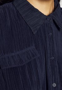 Gros plan sur une chemise plissée bleu marine avec col pointu, devant boutonné et une poche à rabat sur la poitrine.