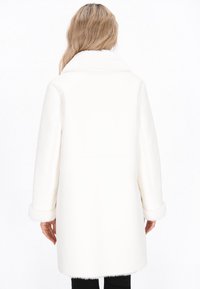Vrouw met lang blond haar, gekleed in een witte middellange jas met een bontkraag en -manchetten, staand met haar rug naar de camera tegen een effen achtergrond.