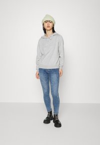 Sweatshirt cinza com capuz e cordão, combinado com jeans skinny azuis e botas pretas de atacadores. O modelo usa um gorro verde claro.