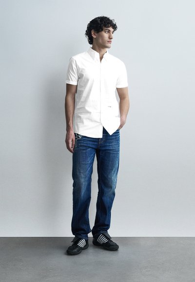 Homme aux cheveux bouclés portant une chemise blanche à manches courtes boutonnée, un jean bleu et des baskets noires à lacets blancs, debout contre un mur gris.