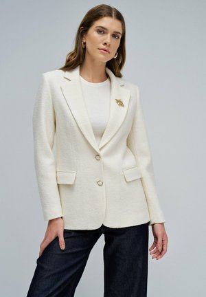 Blazer blanc en tissu texturé, avec un col cranté, deux poches avant, une fermeture à boutons dorés et une broche décorative.