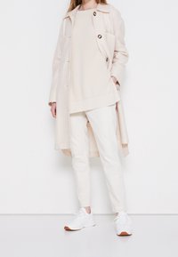 Žena v krémovém dlouhém kabátu, pleteném svetru, úzkých kalhotách a bílých teniskách, stojící na bílém pozadí.