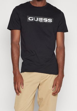 Homme portant un T-shirt noir à manches courtes avec le logo "GUESS" en blanc sur la poitrine et un pantalon beige sur fond uni.