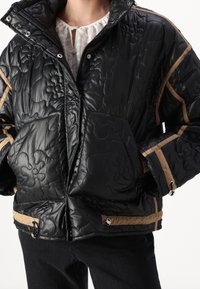 Czarna pikowana kurtka puchowa z kwiecistym wzorem, jasnobrązowymi dodatkami, zatrzaskami oraz dwoma przednimi kieszeniami; wysoki kołnierz i gruba tekstura.