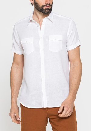 Uomo che indossa una camicia bianca a maniche corte con due tasche sul petto e pantaloni marrone, in piedi davanti a uno sfondo semplice.