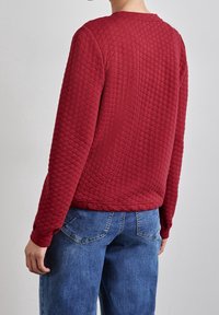 Sudadera acolchada roja con un patrón texturizado y un escote redondo, combinada con unos pantalones vaqueros azules que cuentan con bolsillos traseros.