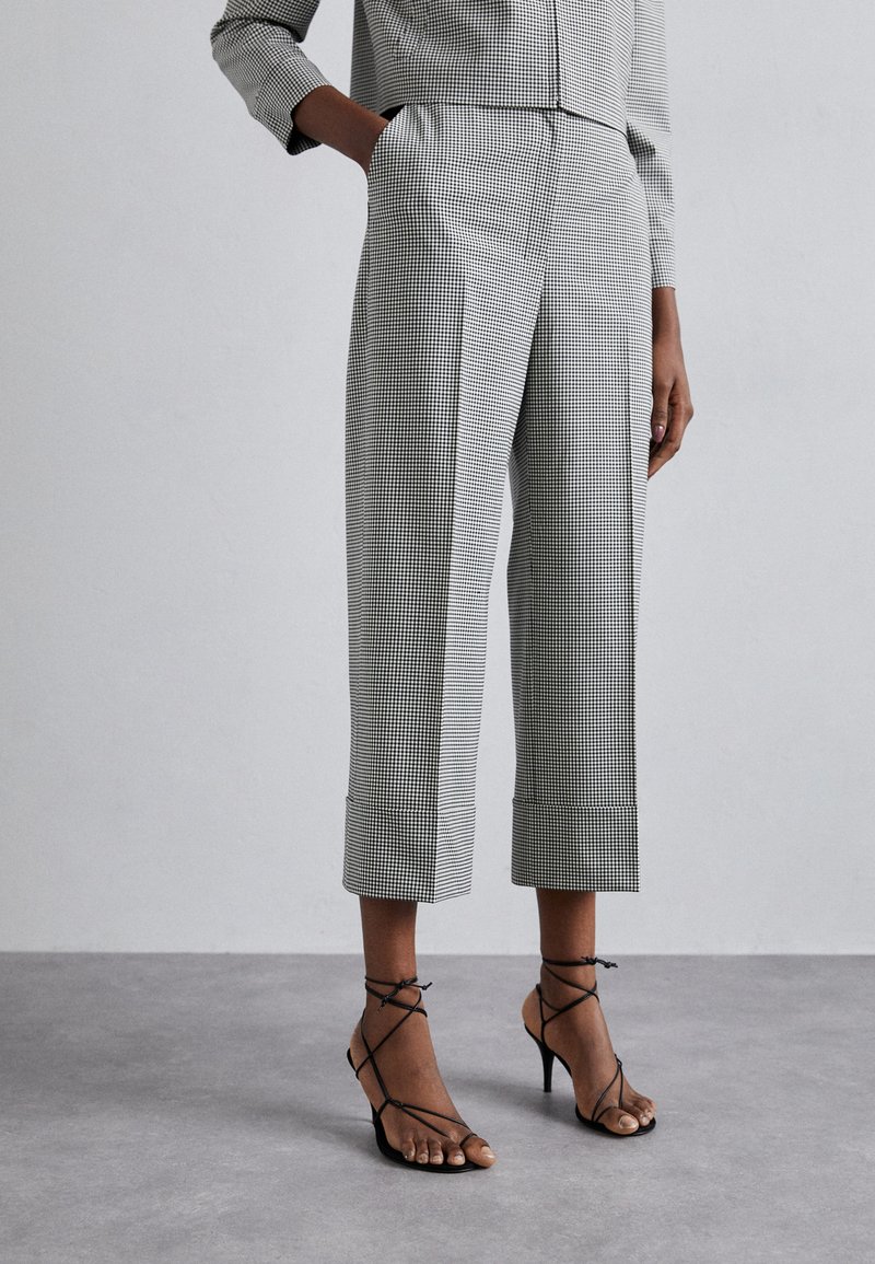 Pantalons cropped en pied de poule noir et blanc avec poches latérales, présentant une coupe ajustée et des ourlets roulés, associés à des talons aiguilles noirs à lanières.
