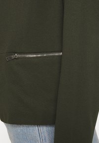 Mörkgrön jacka med en strukturerad tyg, med en silverfärgad ficka med dragkedja som framhäver designen, mot en ljusblå denim.