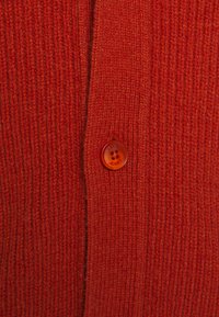 Czerwony kardigan z prążkowanego materiału o fakturowanym splocie, z okrągłym pomarańczowym guzikiem z metką marki, prezentujący prosty i funkcjonalny design.