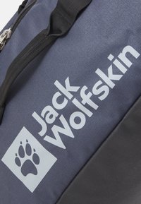 Borsa da viaggio in stoffa blu con dettagli neri, caratterizzata da un logo bianco con la scritta "Jack Wolfskin" e un grafico di zampa all'interno di un contorno quadrato.