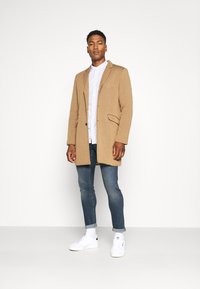 Brown wool blend coat with a notched collar, single button closure, side pockets, worn over a white striped shirt and blue jeans. White sneakers.