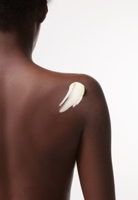 Body lotion på slät, mörk hud. Krämig, off-white lotion applicerad i ett virvlande mönster på axeln. Mjuk textur mot huden.