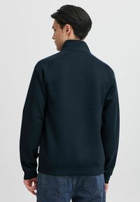 Marinefarget fleecejakke med høy krage og ribbestrikkede mansjetter, med en glatt tekstur og minimalistisk design. Bakviewet viser subtile sømmer og sting.