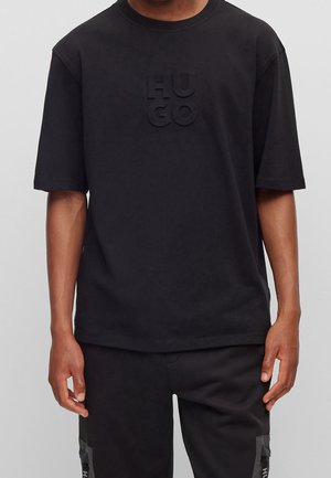 Mężczyzna w czarnym T-shircie z krótkim rękawem z wypukłym logo "HUGO" na klatce piersiowej oraz czarnych spodniach z bocznymi kieszeniami na szarym tle.
