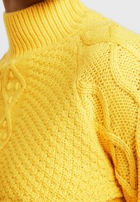 Pull jaune en tricot avec un col montant côtelé, des motifs texturés et des détails en torsades sur les manches et le corps.