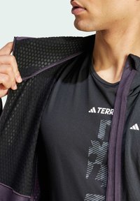 Giacca nera con fodera in rete e accenti viola, indossata sopra una maglietta nera con logo del marchio e design geometrico. Look aderente.