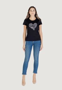 Zwarte T-shirt met een patroon van een hartontwerp, gecombineerd met blauwe jeans en huidskleurige hakken. De stof lijkt glad en heeft een aansluitende vorm.