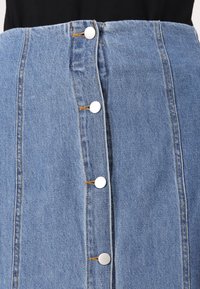 Jupe en denim à taille haute, avec une patte de boutons centrale ornée de boutons blancs et des coutures visibles. De couleur bleu clair avec une texture lisse.