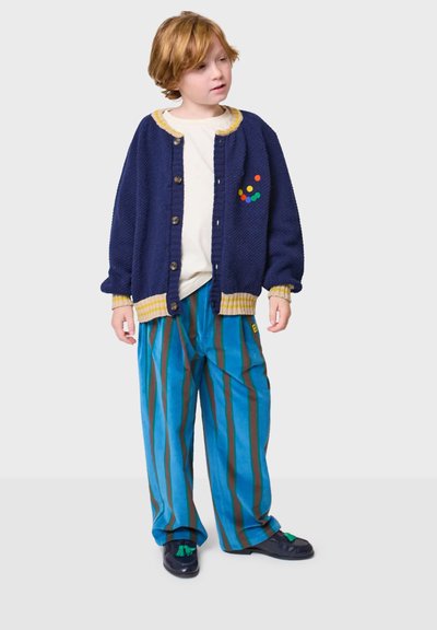 Pleteni tamnoplavi kardigan s žutim obrubom, plave prugaste hlače i crne cipele sa zelenim detaljima; kardigan ima šarene točkice.