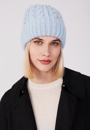 Lichtblauwe gebreide beanie met een gestructureerd kabelpatroon, voorzien van een geribbelde band. Draagt bij een crèmekleurige trui en een zwarte jas.