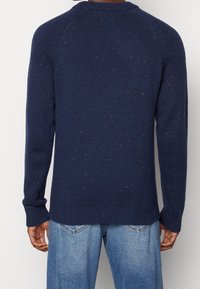 Man som bär en marinblå prickig stickad tröja och ljusblå denimjeans, sedd bakifrån mot en plain bakgrund.