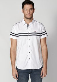 Camisa blanca de manga corta con rayas horizontales negras y un pequeño logo. Diseño con botón y dobladillo redondeado, y cuello en contraste gris.