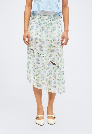 Kobieta nosząca białą spódnicę midi z niebieskim kwiatowym wzorem i ukośnym wycięciem, zestawioną z białymi czółenkami na szpic i jasnoniebieską bluzką.