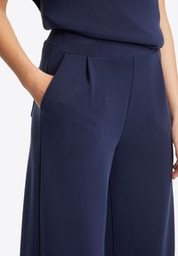 Pantaloni a gamba larga blu navy con una texture liscia, caratterizzati da una parte anteriore plissettata e tasche laterali. Il tessuto sembra leggero e morbido.