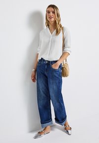 Kvinna lutad mot vit vägg iförd en vit knäppt skjorta, uppvikta blå jeans, silverfärgade ballerinaskor och en beige axelväska.