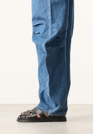 Persoon draagt blauwe denim cargobroek en zwarte leren gevlochten sandalen, staand op een lichte vloer met een effen achtergrond.