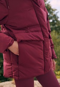 Burgundska puffa jakna s teksturiranim, izoliranim dizajnom. Ima veliki prednji džep i podesivu kapuljaču. Mekan, mat završetak.