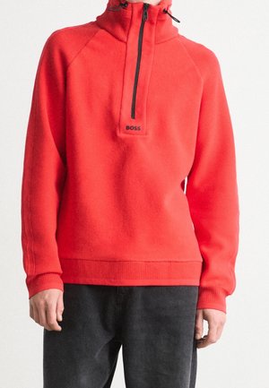 Pull-over en polaire rouge avec une fermeture éclair noire à mi-hauteur sur le devant et un logo "BOSS" sous la fermeture éclair, porté avec un pantalon noir.
