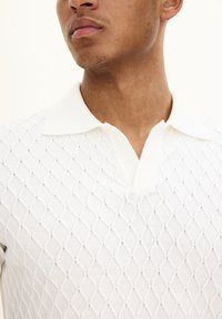 Vit stickad polo med en diamantmönstrad textur, med klassisk krage och korta ärmar, tillverkad av lätt material.