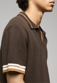 Tricou polo maro tricotat cu finisaj texturat. Are un guler cu dungi și mâneci scurte cu accente albe și maro deschis.