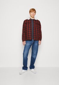 Casaco de flanela xadrez vermelho e preto com colarinho de sheepskin branco, combinado com jeans azuis e ténis brancos. Estilo casual, ajuste relaxado.