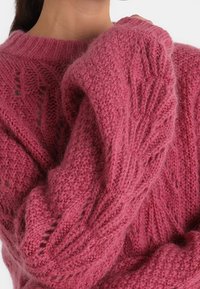 Pull tricoté rose à coupe ample, présentant un motif texturé, un col côtelé et des manches volumineuses. Fabriqué en fil doux et duveteux.