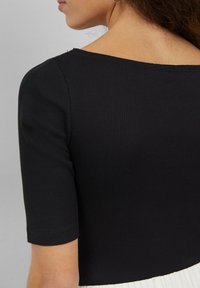 Czarny prążkowany top z szerokim dekoltem i krótkimi rękawami. Materiał ma teksturowany wzór, z widocznymi detalami ściegów przy dolnym brzegu.