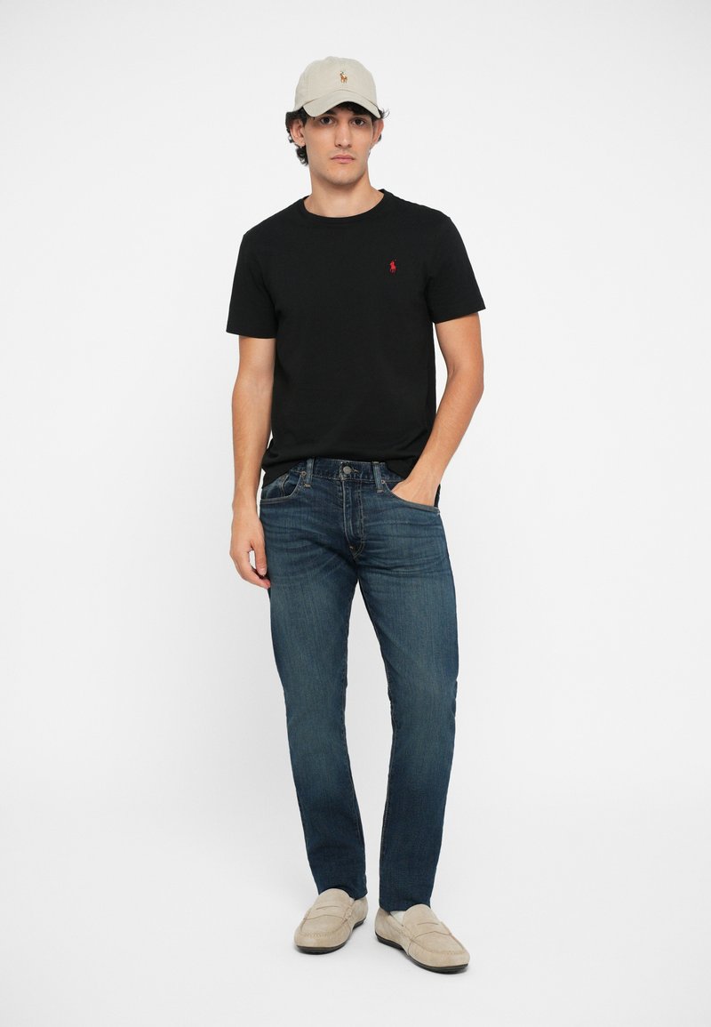 T-shirt noir à manches courtes avec logo rouge, jean en denim bleu, casquette beige et chaussures slip-on beige exposés sur un modèle masculin.