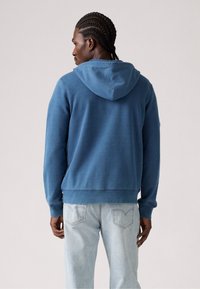 Hoodie in vervaagd blauw, gemaakt van zachte stof. Voorzien van een trekkoord bij de capuchon, lange mouwen en een ribbelrand, gecombineerd met lichtblauwe spijkerbroek.