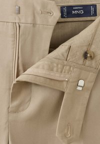 Pantalon beige avec fermeture éclair, crochet métallique, bouton et étiquette MNG Essentials indiquant la taille et l’origine.