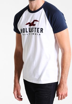Kortärmelig raglan T-shirt in wit met marineblauwe mouwen. Voorzien van het "HOLLISTER CALIFORNIA" logo en een adelaar afbeelding in bordeauxrood. Materiaal van katoen.